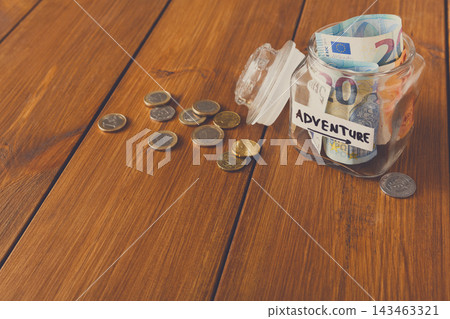 A clear jar is filled with banknotes and coins on a wooden table. The jar has a label that says adventure. Some coins are spilled out next to the jar, suggesting saving for future travel. 143463321