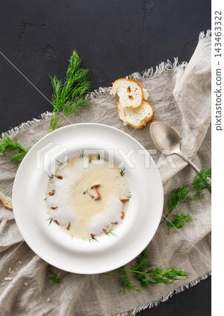 A bowl of soup sits on a neutral linen cloth. The soup has bits of herbs and is accompanied by slices of bread. A metal spoon rests nearby, creating a simple and inviting meal setting. A bowl of soup sits on a neutral linen cloth. The soup has bits of herbs and is accompanied by slices of bread. A metal spoon rests nearby, creating a simple and inviting meal setting. 143463322