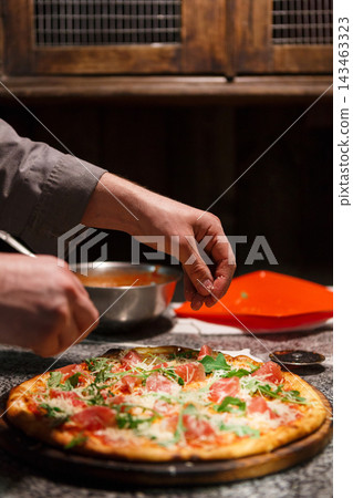 A person is preparing a pizza by adding toppings in a cozy kitchen. Fresh ingredients like tomatoes and herbs are being sprinkled on top. The setting is warm and inviting. 143463323