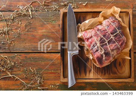 A cut of meat is placed on a wooden board next to a sharp knife. The setting includes a rustic wooden table with hay scattered around, showing preparation for cooking. A cut of meat is placed on a wooden board next to a sharp knife. The setting includes a rustic wooden table with hay scattered around, showing preparation for cooking. 143463399