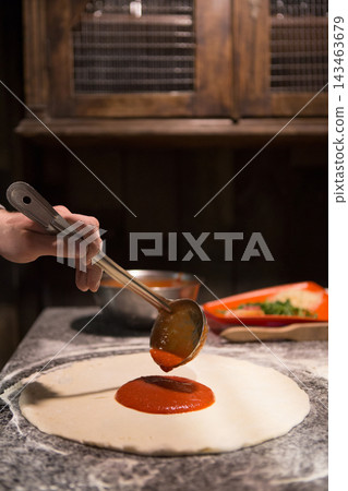 A person pours tomato sauce onto pizza dough on a countertop in a kitchen. Other ingredients are placed nearby. This scene takes place during the evening. A person pours tomato sauce onto pizza dough on a countertop in a kitchen. Other ingredients are placed nearby. This scene takes place during the evening. 143463679