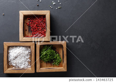 Three wooden containers hold different spices. One contains pink peppercorns, another has white salt, and the last holds fresh herbs. The setting has a dark surface that highlights the spices. Three wooden containers hold different spices. One contains pink peppercorns, another has white salt, and the last holds fresh herbs. The setting has a dark surface that highlights the spices. 143463725