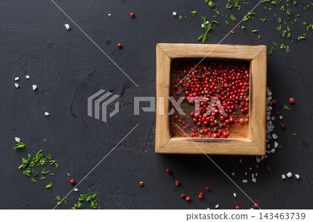 Red peppercorns fill a wooden square container. Salt and fresh herbs are scattered on a black countertop. This setup shows elements typical in a kitchen during meal preparation. Red peppercorns fill a wooden square container. Salt and fresh herbs are scattered on a black countertop. This setup shows elements typical in a kitchen during meal preparation. 143463739