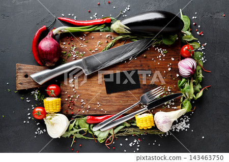 Fresh vegetables like eggplant, chili, and onion are placed on a wooden cutting board. A knife and fork are included with salt and herbs around the board. Colorful ingredients are ready for cooking. Fresh vegetables like eggplant, chili, and onion are placed on a wooden cutting board. A knife and fork are included with salt and herbs around the board. Colorful ingredients are ready for cooking. 143463750