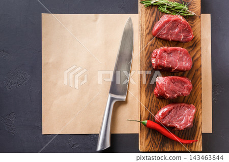 Four cuts of raw beef are arranged on a wooden board next to a sharp knife. Fresh herbs and a red chili are also placed nearby. The setting is a kitchen counter and paper is underneath. Four cuts of raw beef are arranged on a wooden board next to a sharp knife. Fresh herbs and a red chili are also placed nearby. The setting is a kitchen counter and paper is underneath. 143463844