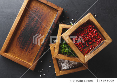 Three wooden containers are arranged on a dark surface. One holds pink peppercorns, another has green herbs, and the third contains salt crystals. This scene shows a culinary setting. Three wooden containers are arranged on a dark surface. One holds pink peppercorns, another has green herbs, and the third contains salt crystals. This scene shows a culinary setting. 143463996