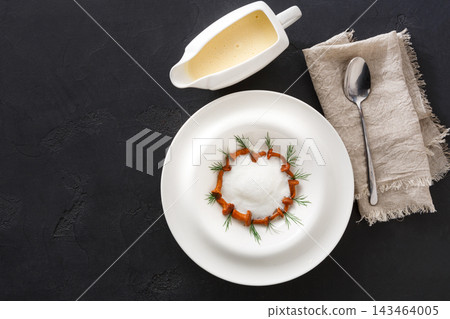 A dish is arranged in a circular pattern with cream and garnished with herbs. A side of sauce is presented in a small dish. A spoon and cloth napkin are nearby. A dish is arranged in a circular pattern with cream and garnished with herbs. A side of sauce is presented in a small dish. A spoon and cloth napkin are nearby. 143464005