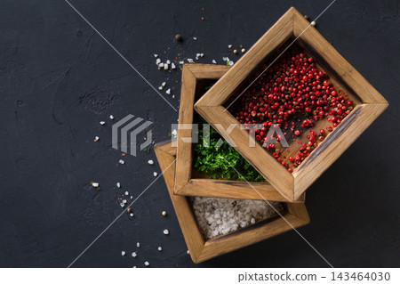 Three wooden boxes are stacked on a dark surface. One box contains red peppercorns, another has herbs, and the last holds salt. They are staged for cooking preparation. Three wooden boxes are stacked on a dark surface. One box contains red peppercorns, another has herbs, and the last holds salt. They are staged for cooking preparation. 143464030