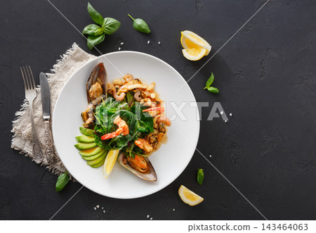 The plate shows a mix of shrimp, clams, and greens. Slices of lemon and avocado are arranged on the side. The setting is simple with a dark background and cutlery. The plate shows a mix of shrimp, clams, and greens. Slices of lemon and avocado are arranged on the side. The setting is simple with a dark background and cutlery. 143464063