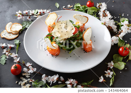 A white plate holds a salad featuring salmon wrapped with greens and topped with cheese. Cherry tomatoes and herbs surround the plate, enhancing the dish's look on a dark table. A white plate holds a salad featuring salmon wrapped with greens and topped with cheese. Cherry tomatoes and herbs surround the plate, enhancing the dish's look on a dark table. 143464064