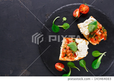 Two pieces of bread topped with tomato, cheese, and greens are placed on a black plate. Cherry tomatoes and fresh leaves surround the bread, showcasing a casual dining setting. Two pieces of bread topped with tomato, cheese, and greens are placed on a black plate. Cherry tomatoes and fresh leaves surround the bread, showcasing a casual dining setting. 143464083