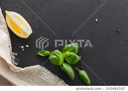A piece of lemon rests beside fresh basil leaves on a dark surface. Crystals of salt are scattered nearby, along with a piece of cloth adding texture to the setting. A piece of lemon rests beside fresh basil leaves on a dark surface. Crystals of salt are scattered nearby, along with a piece of cloth adding texture to the setting. 143464165