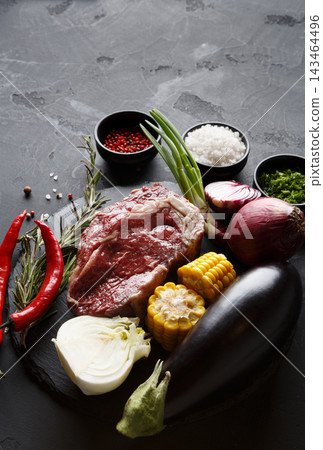 Fresh ingredients are arranged on a dark surface. The setup includes meat, corn, eggplant, onions, green onions, and spices. This collection is ready for cooking a flavorful meal. Fresh ingredients are arranged on a dark surface. The setup includes meat, corn, eggplant, onions, green onions, and spices. This collection is ready for cooking a flavorful meal. 143464496