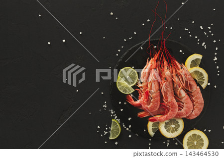 Shrimp are arranged on a black plate with lemon slices placed around them. Sea salt is sprinkled nearby. This setup is meant for serving seafood. Shrimp are arranged on a black plate with lemon slices placed around them. Sea salt is sprinkled nearby. This setup is meant for serving seafood. 143464530