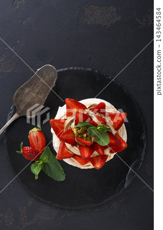 A dessert sits on a dark surface. It is topped with fresh strawberries and mint leaves. A serving spoon rests beside the dessert, ready for serving. The setting is simple and clean. A dessert sits on a dark surface. It is topped with fresh strawberries and mint leaves. A serving spoon rests beside the dessert, ready for serving. The setting is simple and clean. 143464584