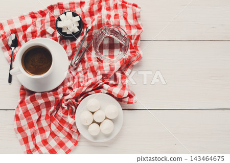 A cup of coffee sits on a white plate next to a bowl of milk sweets and a dish of sugar cubes. A glass of water is present on a red and white checkered cloth. A cup of coffee sits on a white plate next to a bowl of milk sweets and a dish of sugar cubes. A glass of water is present on a red and white checkered cloth. 143464675