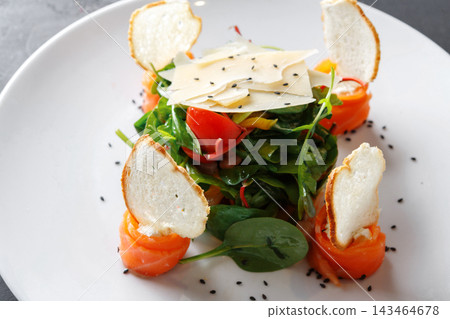 A salad features greens topped with slices of smoked salmon and crispy bread. Red tomatoes and cheese are visible. The dish has black seeds sprinkled around it and is served on a white plate. 143464678