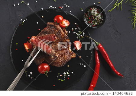 A grilled steak rests on a black plate surrounded by cherry tomatoes, spices, and chili peppers. A fork is ready to slice the meat. The setting is simple with a dark table. A grilled steak rests on a black plate surrounded by cherry tomatoes, spices, and chili peppers. A fork is ready to slice the meat. The setting is simple with a dark table. 143464682