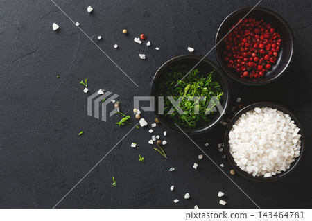 Three bowls are placed on a dark surface. One bowl contains salt, another holds red peppercorns, and the third has chopped herbs, creating a simple setup for cooking. Three bowls are placed on a dark surface. One bowl contains salt, another holds red peppercorns, and the third has chopped herbs, creating a simple setup for cooking. 143464781