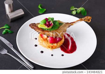A duck leg is placed on a mound of rice beside a sauce made with fruit. There are greens and garnishes on top. The setting shows a simple dining scene. 143464821