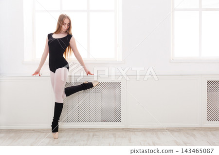 A dancer in a black and gray outfit practices ballet in a bright studio. She stands on one leg, balancing with grace near a white wall. Sunlight fills the space through large windows. A dancer in a black and gray outfit practices ballet in a bright studio. She stands on one leg, balancing with grace near a white wall. Sunlight fills the space through large windows. 143465087