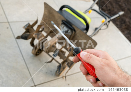 In an outdoor setting, a technician holds a screwdriver for repairing a garden tiller machine. The technician is preparing tools to fix the tillers components on a concrete surface. In an outdoor setting, a technician holds a screwdriver for repairing a garden tiller machine. The technician is preparing tools to fix the tillers components on a concrete surface. 143465088