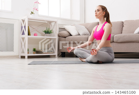 A woman is sitting cross-legged on a yoga mat in a living room. She has her eyes closed and is focused on breathing. Natural light comes from the windows. A couch and a shelf are in the background. A woman is sitting cross-legged on a yoga mat in a living room. She has her eyes closed and is focused on breathing. Natural light comes from the windows. A couch and a shelf are in the background. 143465100