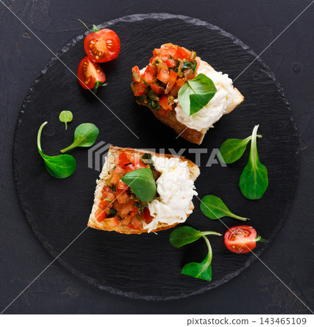 Two pieces of bruschetta are placed on a black plate. One is topped with tomato mix and basil, while the other has cheese. Cherry tomatoes and greens surround the bruschetta. Two pieces of bruschetta are placed on a black plate. One is topped with tomato mix and basil, while the other has cheese. Cherry tomatoes and greens surround the bruschetta. 143465109