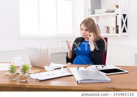 A pregnant woman sits at her desk in an office. She speaks on the phone with her left hand raised. Documents, a laptop, and plants are on the desk. The room has bright light coming from a window. A pregnant woman sits at her desk in an office. She speaks on the phone with her left hand raised. Documents, a laptop, and plants are on the desk. The room has bright light coming from a window. 143465110