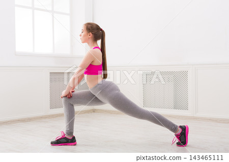 A young woman performs a stretch in a room with natural light. She is wearing a sports outfit and has her hair tied back. The space appears clean and open, ideal for exercising. A young woman performs a stretch in a room with natural light. She is wearing a sports outfit and has her hair tied back. The space appears clean and open, ideal for exercising. 143465111