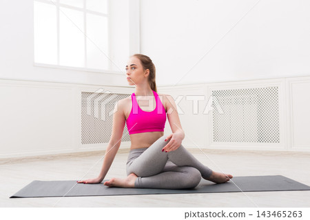 A young woman practices yoga on a mat in a bright studio. She sits with one leg crossed over the other. Sunlight enters through the window, creating a warm atmosphere for exercise. 143465263