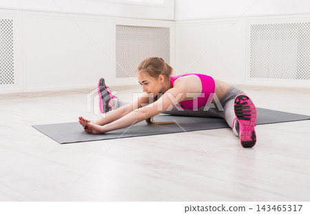 A young woman performs a stretching exercise on a yoga mat in a bright room with wooden flooring. She focuses on her movements while wearing sportswear. A young woman performs a stretching exercise on a yoga mat in a bright room with wooden flooring. She focuses on her movements while wearing sportswear. 143465317