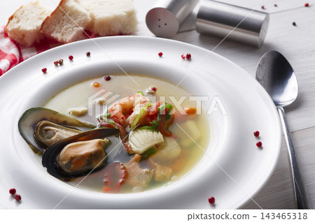 A bowl of seafood soup contains shrimp, mussels, and vegetables. The dish is placed on a table with a spoon, salt shaker, bread, and scattered pepper. A bowl of seafood soup contains shrimp, mussels, and vegetables. The dish is placed on a table with a spoon, salt shaker, bread, and scattered pepper. 143465318