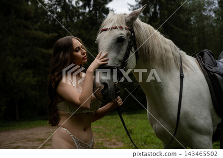 Woman in lace lingerie stroking a white horse in a clearing Woman in lace lingerie stroking a white horse in a clearing 143465502