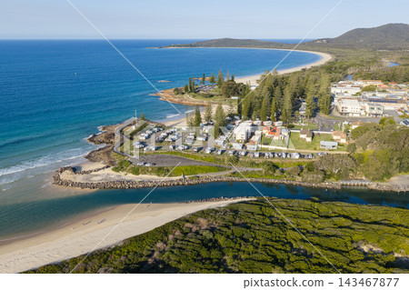 Aerial view across Back Creek and its sandy banks toward a coastal campground, rocky headlands and blue ocean. Tall trees border the waterfront, while a long curved beach stretches toward wooded hills Aerial view across Back Creek and its sandy banks toward a coastal campground, rocky headlands and blue ocean. Tall trees border the waterfront, while a long curved beach stretches toward wooded hills 143467877