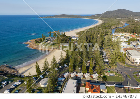 Aerial view of Horseshoe Beach beside a campground shaded by tall trees, with Front Beach curving around the bay in the background. Rocky headlands, waterfront buildings and coastal woodland border 143467878