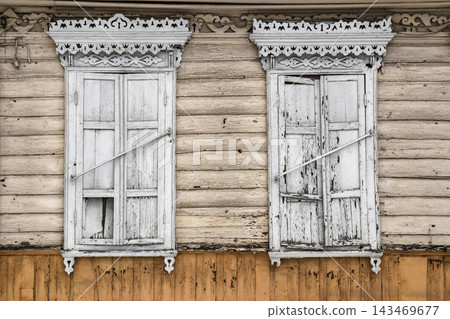 Two white windows on a wooden house show signs of age. The walls are made of light wood, and the windows have peeling paint and broken frames 143469677