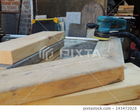 A blue random orbital sander rests on a workbench next to a stack of wooden boards and blocks. A close-up view of the process of preparing, sanding, and leveling wood in a carpentry workshop. A blue random orbital sander rests on a workbench next to a stack of wooden boards and blocks. A close-up view of the process of preparing, sanding, and leveling wood in a carpentry workshop. 143472028
