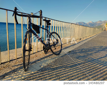 Dark Blue Triban Road Bike Leaning against Metal Fence on Coastal Promenade at Sunset. Dark Blue Triban Road Bike Leaning against Metal Fence on Coastal Promenade at Sunset. 143480207