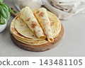 Stack of lavash or chapati flatbreads on a wooden cutting board, with a jar of flour, fresh basil 143481105