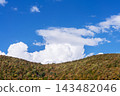 Large white clouds above wooded autumn hills beneath blue sky. Mountain landscape and cloudscape. Weather and atmospheric conditions. Nature, environment and seasonal scenery. 143482046