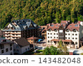 Mountain village buildings below a densely forested hillside. Alpine architecture and wooded slope. Mountain settlement, European village landscape. Built environment and nature 143482048
