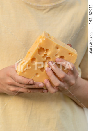 Person's hands carefully holding a large, golden block of dairy cheese with distinctive holes, showcasing a healthy ingredient and emphasizing quality food preparation and natural nutrition 143482053
