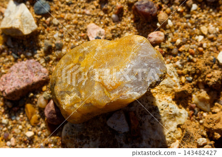 Glossy golden vein quartz pebble stained by iron oxides lies on rough quarry sand, glittering brightly under harsh sun rays Glossy golden vein quartz pebble stained by iron oxides lies on rough quarry sand, glittering brightly under harsh sun rays 143482427