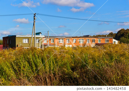 Ordinary low-rise apartment building in provincial Russian countryside behind wild grass thickets under blue sky Ordinary low-rise apartment building in provincial Russian countryside behind wild grass thickets under blue sky 143482458