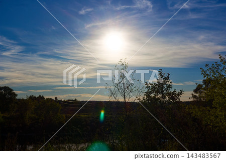 Radiant sun shining brightly above lush green trees in a serene summer landscape, with soft wispy clouds drifting across a deep blue sky during a peaceful golden hour moment Radiant sun shining brightly above lush green trees in a serene summer landscape, with soft wispy clouds drifting across a deep blue sky during a peaceful golden hour moment 143483567