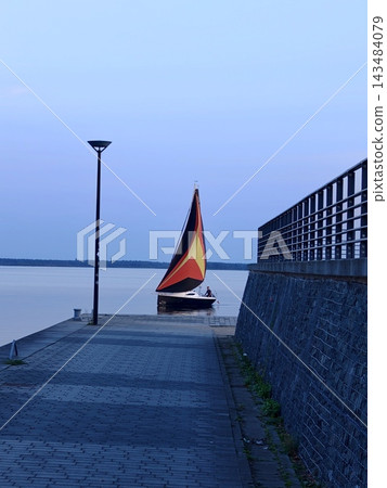 A sailboat with a bright sail approaches the waterfront at sunset. A sailboat with a bright sail approaches the waterfront at sunset. 143484079