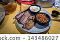 Smoked brisket served with baked beans, creamy mashed potatoes and toast on a casual barbecue plate. Hearty comfort-food meal showcasing tender meat, smoky flavors 143486027