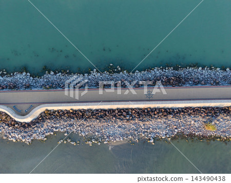 Lake Balaton shore features stone breakwater and paved walkway viewed from overhead. Clear blue green water borders rocks, while nautical emblem marks lakeside promenade near rocky waterline. Lake Balaton shore features stone breakwater and paved walkway viewed from overhead. Clear blue green water borders rocks, while nautical emblem marks lakeside promenade near rocky waterline. 143490438