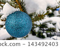 A Christmas tree stands outside covered in snow, with a blue ornament hanging from its branches. The scene shows winter festivities and holiday spirit 143492403